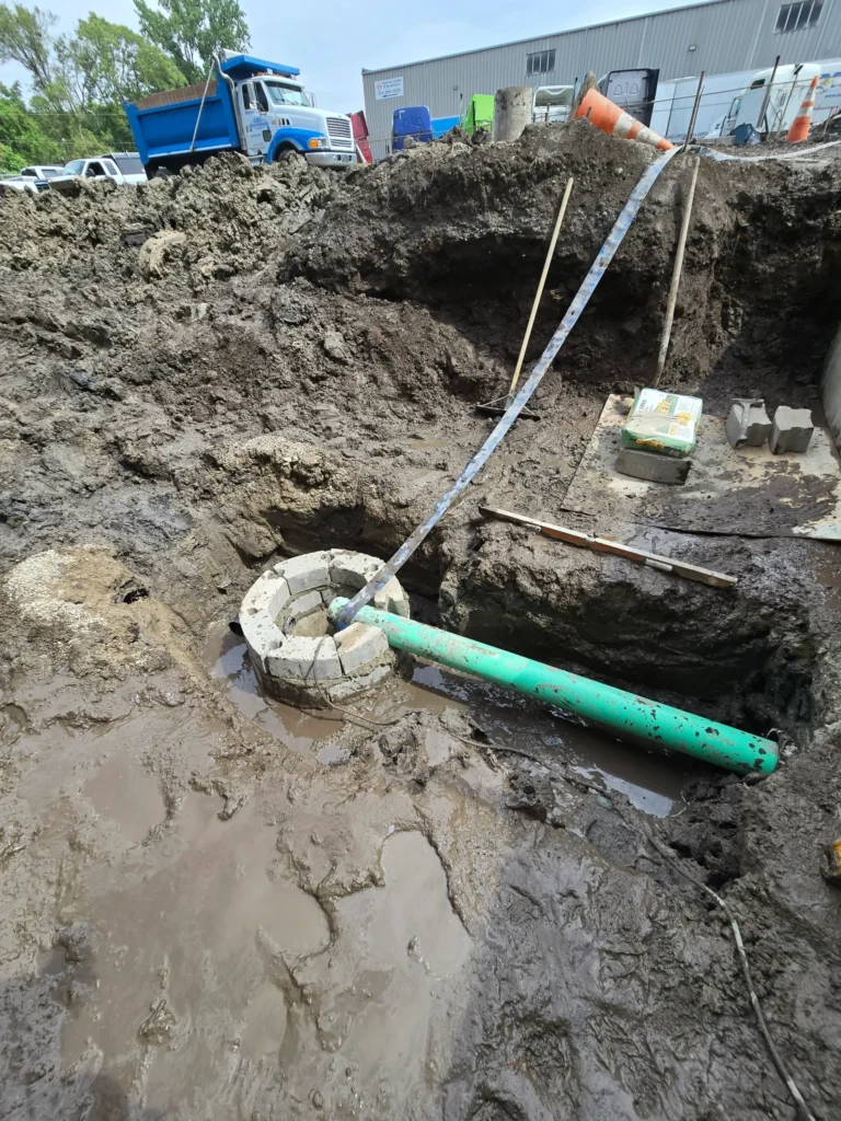 Muddy construction excavation site with a green PVC pipe leading into a circular concrete block structure.