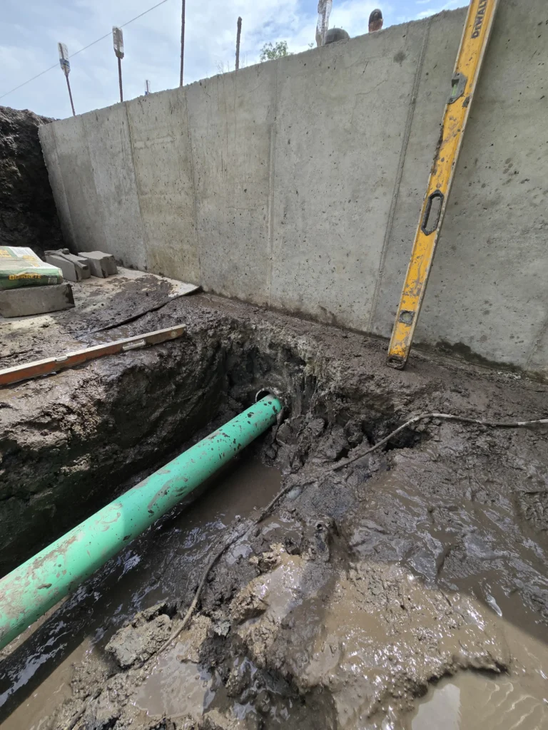 Green pipe in a muddy trench with standing water, next to a concrete wall, with a yellow level and Quikrete bags.