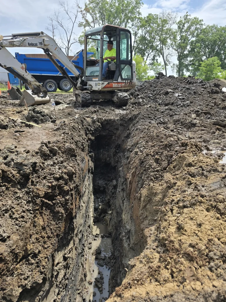 Worker operating a Bobcat excavator, digging a deep, muddy trench at a construction site with dirt piles and a dump truck.