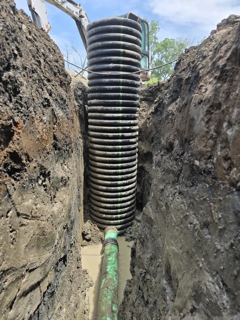 Construction trench with a large corrugated pipe, green pipe in muddy water, and excavator arm visible above.