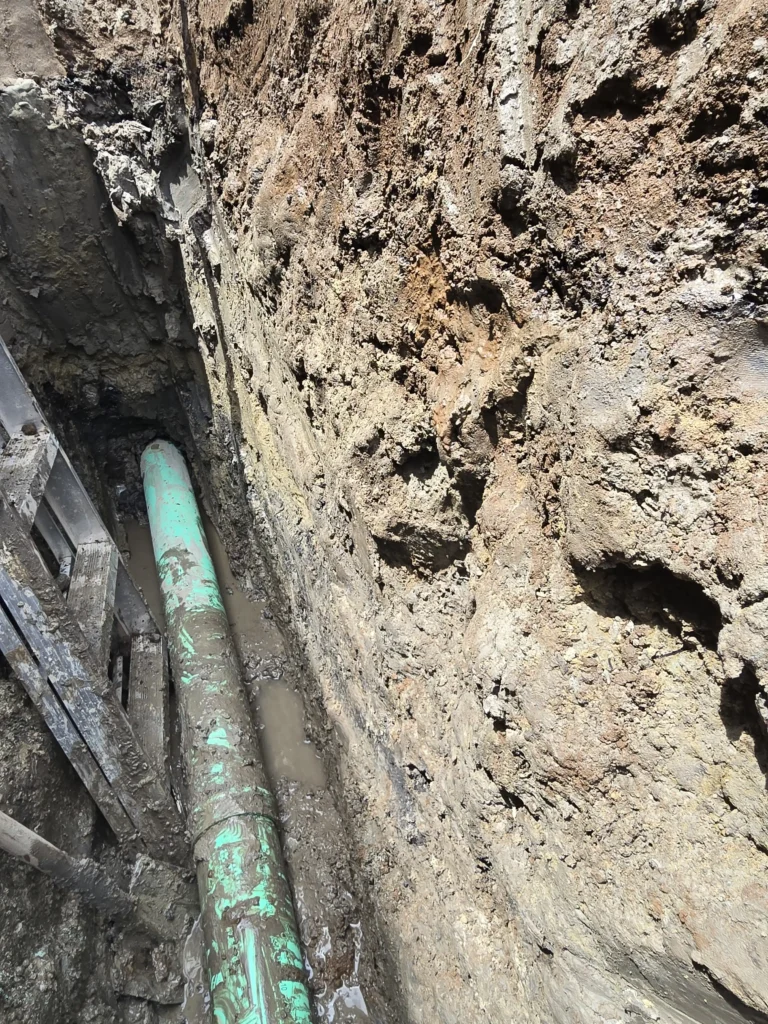 Green pipe laid in a deep, muddy excavation trench with temporary wooden shoring and standing water; rough, layered earth walls.