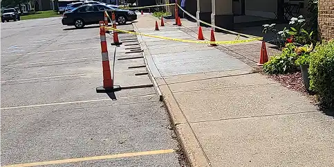 Orange traffic cones and yellow caution tape block a sidewalk adjacent to a parking lot with parked cars and a building.