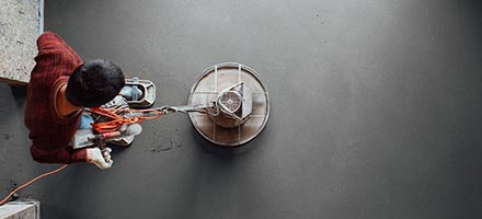 Person in a red shirt and gloves operating a large, round floor polisher on a dark gray concrete floor, viewed from directly above.