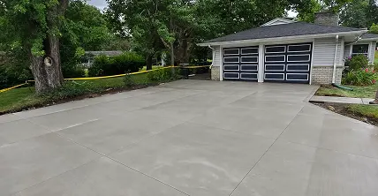 Newly poured concrete driveway leading to a house with light siding and two dark, modern garage doors. Green trees and grass frame the drive, with yellow caution tape along the left lawn.