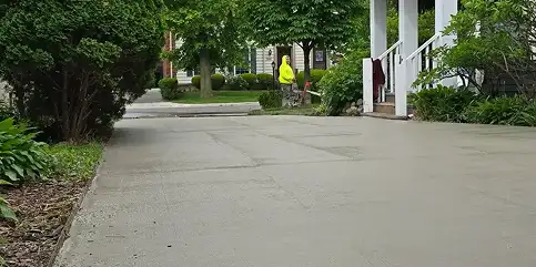 Smooth, new gray concrete driveway with control joints. A person in a yellow jacket uses lawn equipment by the street, a white house porch is on the right.