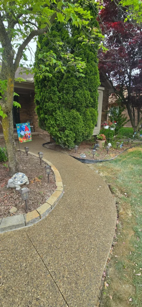 Curved aggregate concrete walkway leading to a house entrance with landscaping, gnome flag, and garden lights.