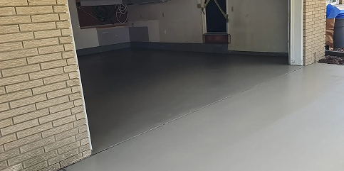 Garage with open door, revealing a dark gray floor inside and a light gray concrete driveway. The house exterior is light brick, and the garage interior features wall-mounted cabinets and a door with yellow caution tape.