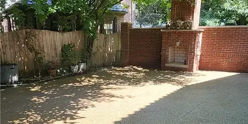 Sunny outdoor patio with light-colored pavement, a wooden fence on the left, and a brick wall with an outdoor fireplace on the right. Tree shadows dappling the ground.
