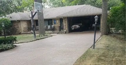 Single-story ranch house with a light-colored driveway leading to an open two-car garage revealing a dark Jeep. A basketball hoop, lamppost, and mature trees are in the front yard.