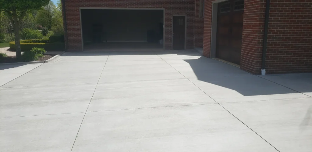 Newly laid light gray concrete driveway in front of a red brick house featuring an open garage, a closed wooden garage, and a dark side door. A landscaped garden with trees and hedges is visible on the left.