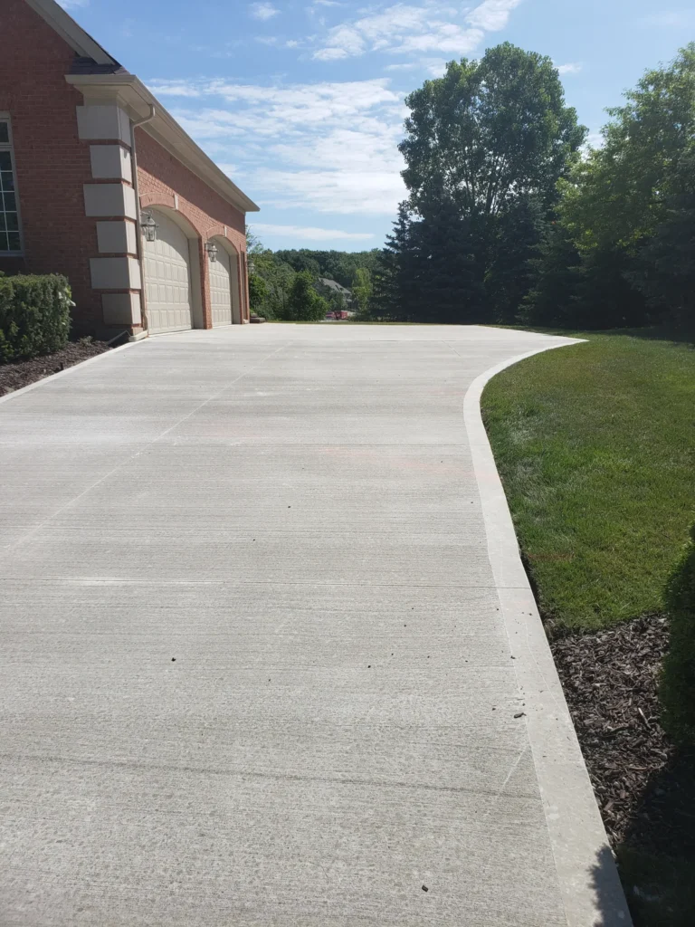 A wide concrete driveway curves past a brick house with two garage doors, bordered by green grass and trees under a bright blue sky with scattered white clouds.