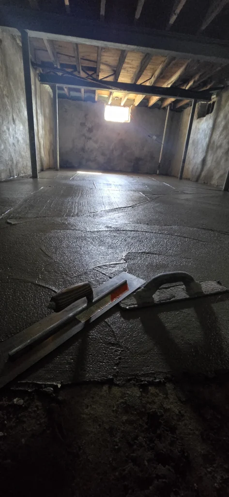 Basement floor freshly poured with wet concrete, with two finishing trowels in the foreground. Rough walls, exposed wooden ceiling joists, and a bright window at the back.