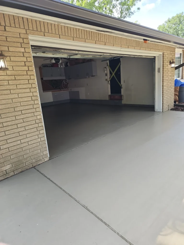 Garage with open door, revealing a dark gray floor inside and a light gray concrete driveway. The house exterior is light brick, and the garage interior features wall-mounted cabinets and a door with yellow caution tape.