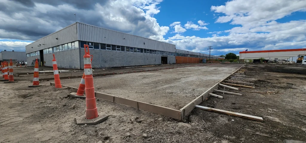 Ground prepared with wooden forms for concrete, orange cones, and a large industrial building under a partly cloudy sky.
