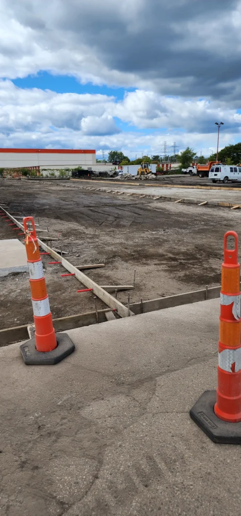 Construction site with two orange and white cones in the foreground, excavated dirt with concrete forms, a building, and construction vehicles in the background under a cloudy sky.