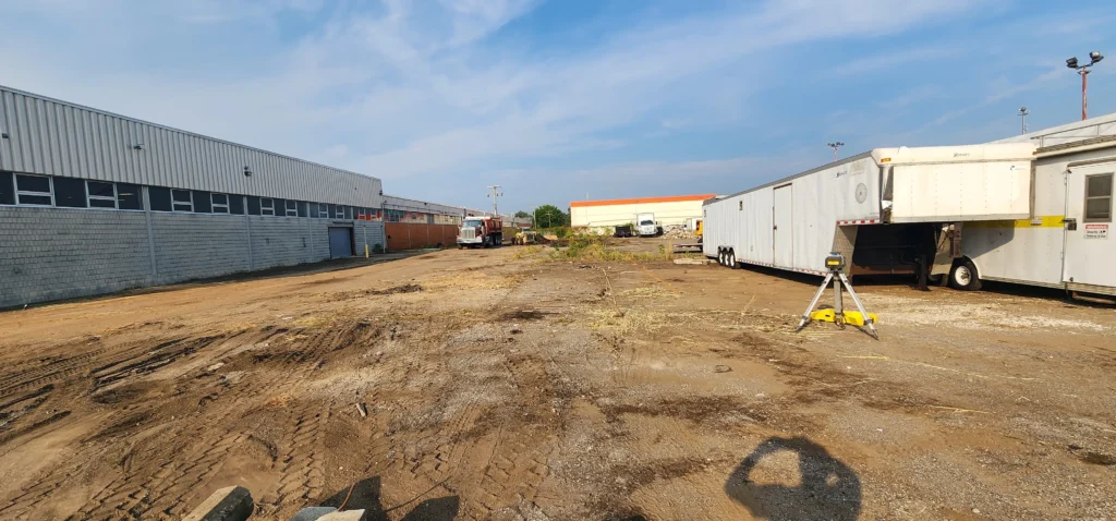 Unpaved industrial lot with tire tracks, bordered by warehouses. A dump truck, excavator, and large white trailer are visible, with a surveying tripod in the foreground.