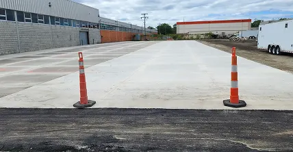 Newly paved concrete lot between industrial warehouse buildings with two orange safety cones in the foreground.