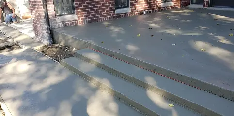 Newly poured concrete patio and three steps with thin red lines embedded in their edges, leading up to a red brick building. Scattered leaves and tree shadows are on the concrete surface. A person is partially visible working near the building's corner.