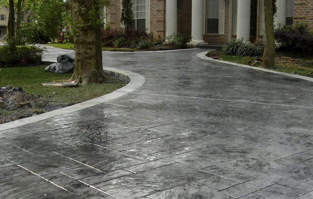 Shiny, dark gray stamped concrete driveway with a rectangular pattern, curving towards a brick house with white columns. Green grass and trees border the drive.