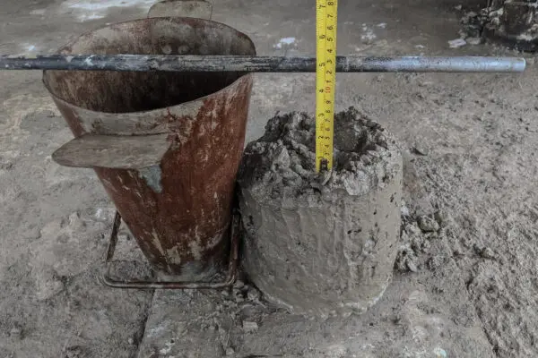 A slump cone positioned next to a pile of concrete that's just been formed with the cone. There is a tape measure in the center of the formed concrete to measure its vertical distance. 