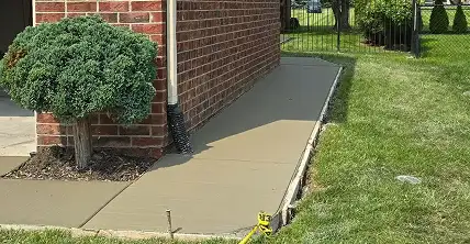 Freshly poured concrete sidewalk next to a brick building, with a green bush and grassy lawn.