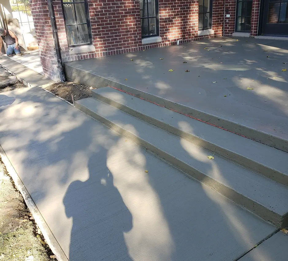 Newly poured concrete patio and three steps with thin red lines embedded in their edges, leading up to a red brick building. Scattered leaves and tree shadows are on the concrete surface. A person is partially visible working near the building's corner.